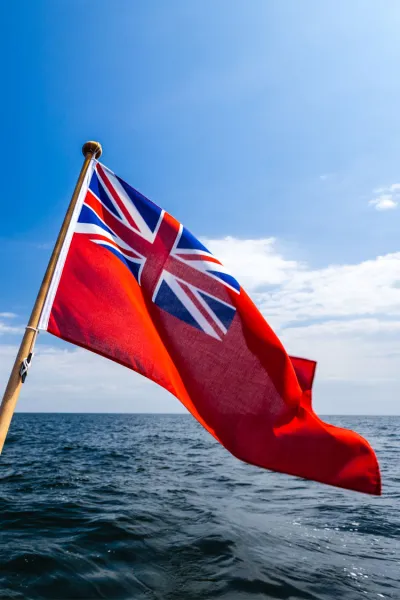 Red ensign flag flies at the back of a boat, with blue skies and open seas in the background