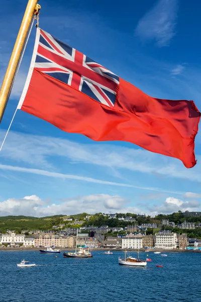 Red Ensign flying in the wind with blue skies, buildings and a variety of vessels in the background