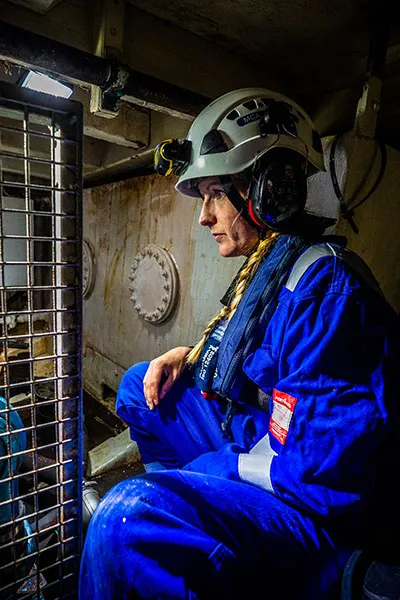 A female maritime surveyor is crouched inside a vessel while wearing a blue boiler suit, lifejacket and a hardhat
