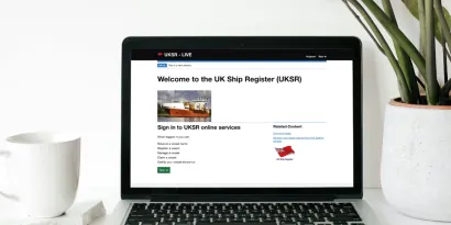 UK Ship Register Online Service web page on a laptop with a coffee cup to the left and a plant to the right