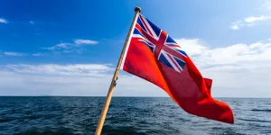 Red ensign flag flies at the back of a boat, with blue skies and open seas in the background