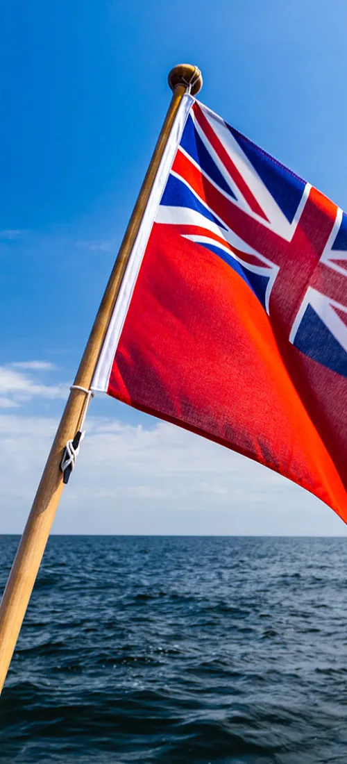 Red ensign flag flies at the back of a boat, with blue skies and open seas in the background